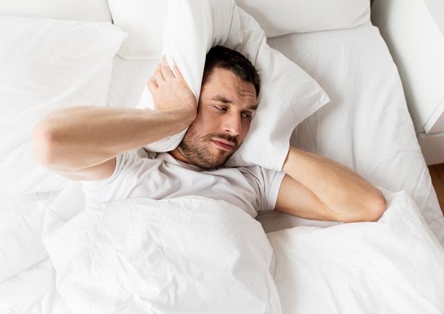 person-laying-in-white-bed-using-a-pillow-to-cover-their-ears
