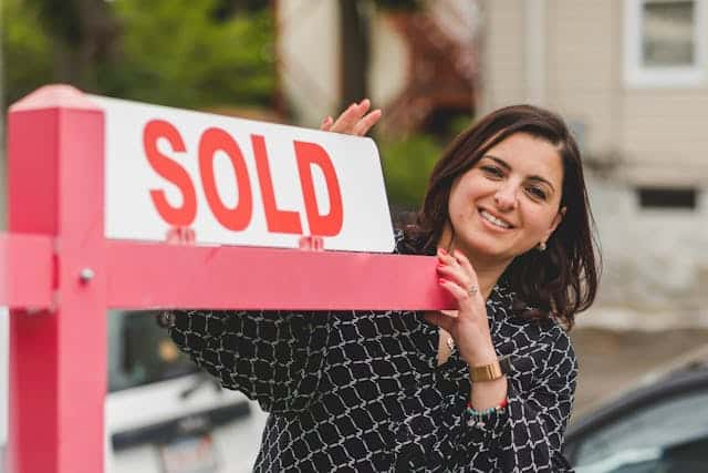 Woman smiling next to a "SOLD" sign in front of a house, indicating a successful real estate transaction.