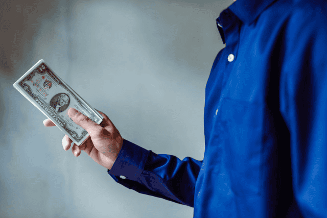 a-person-wearing-a-blue-shirt-holds-a-stack-of-u-s-dollar-bills-in-one-hand-with-a-neutral-background